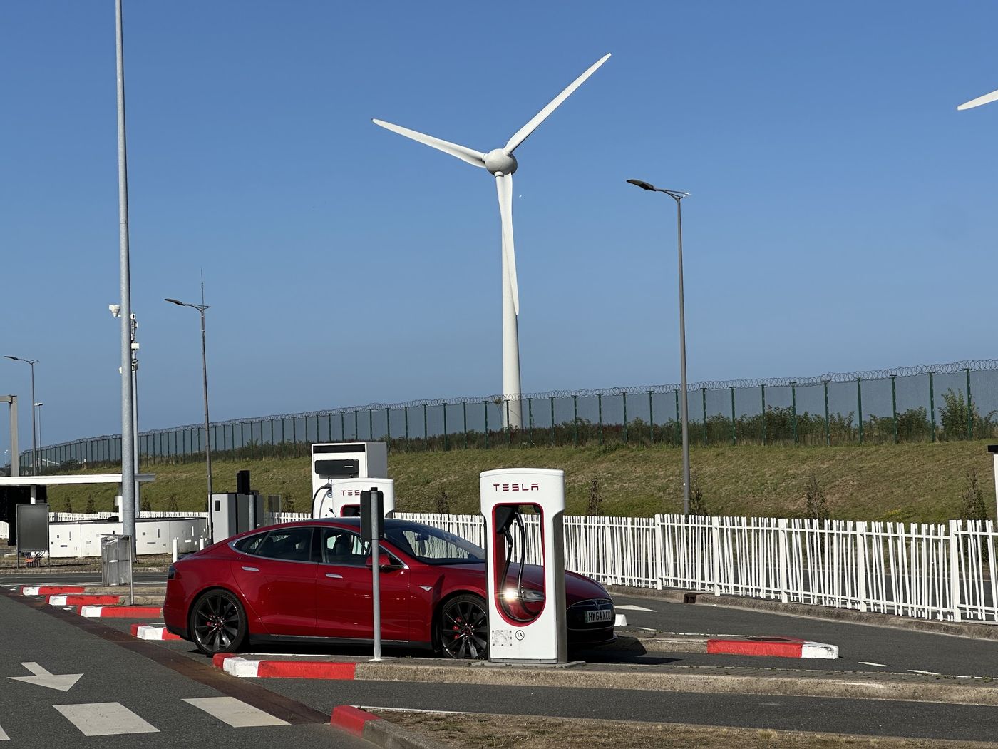 An electric car charging at a rapid charger with a wind turbine directly behind it.