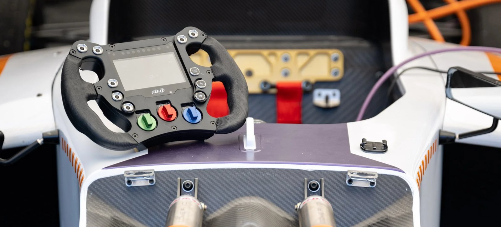 Front suspension and exposed carbon monocoque of an electric formula car, steering wheel and high-voltage cabling visible.