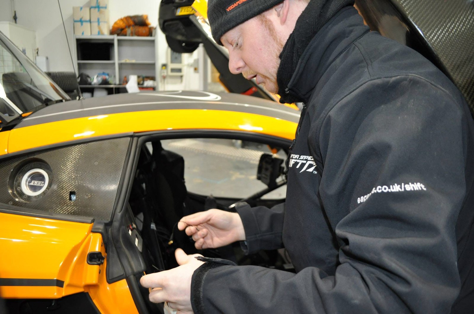 Wiring capture equipment into the cockpit of an orange and carbon race car, wearing a Need for Speed SHIFT jacket.