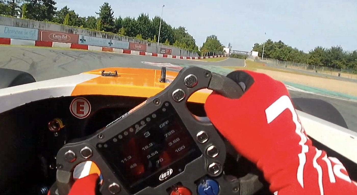 Driver's-eye view from the cockpit of an electric formula car, hands on the wheel with a digital dash mid-corner.