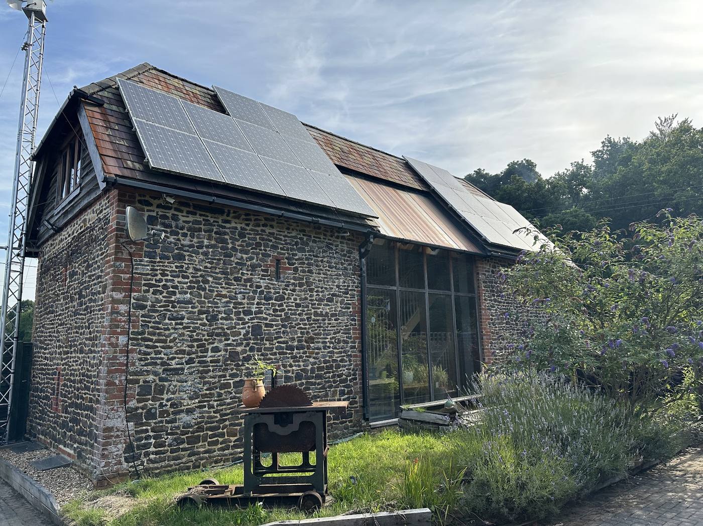 A converted flint barn with solar panels across its roof and a wind turbine mast alongside.